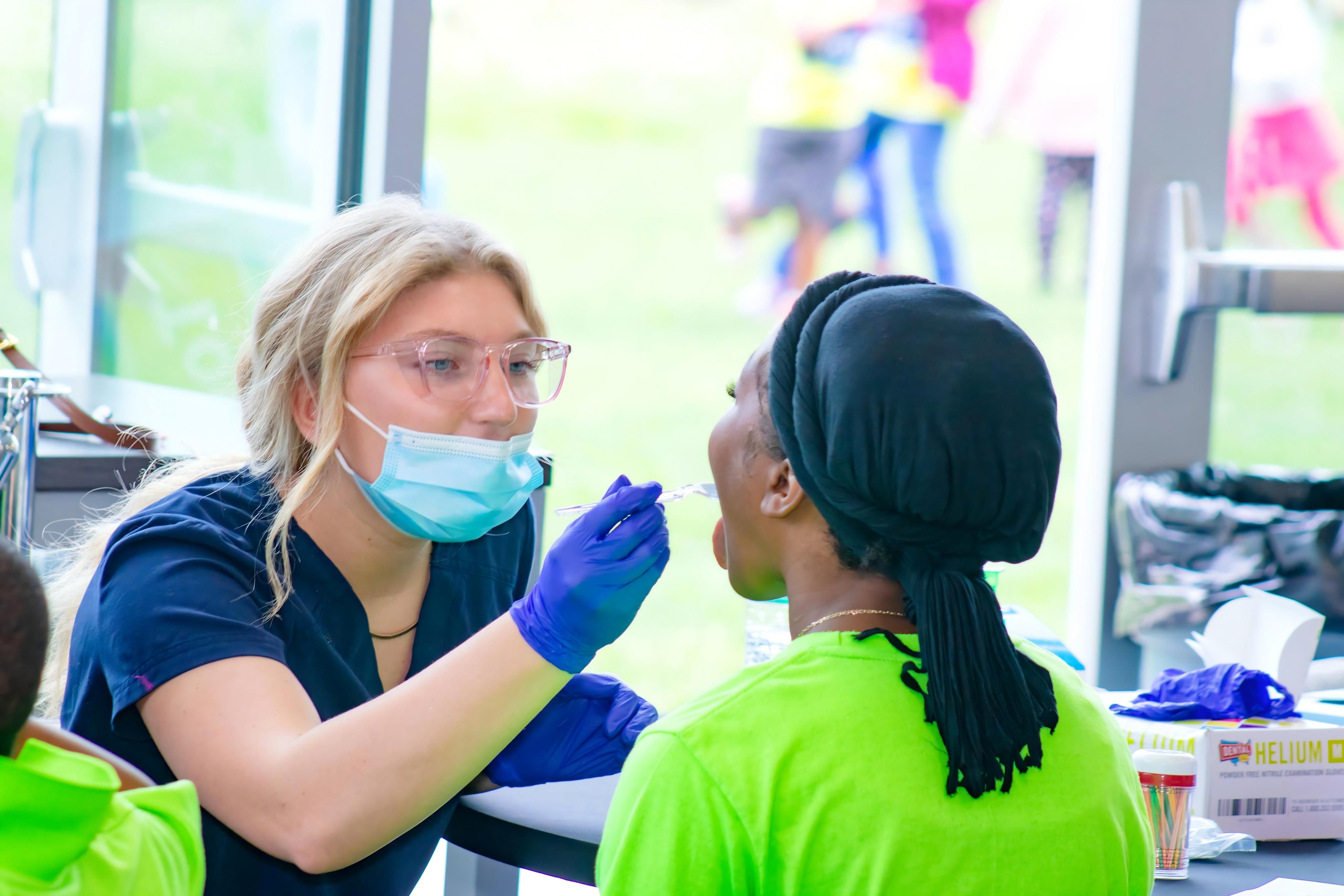 A masked healthcare worker in blue gloves uses a swab near a seated person’s mouth at a screening table.