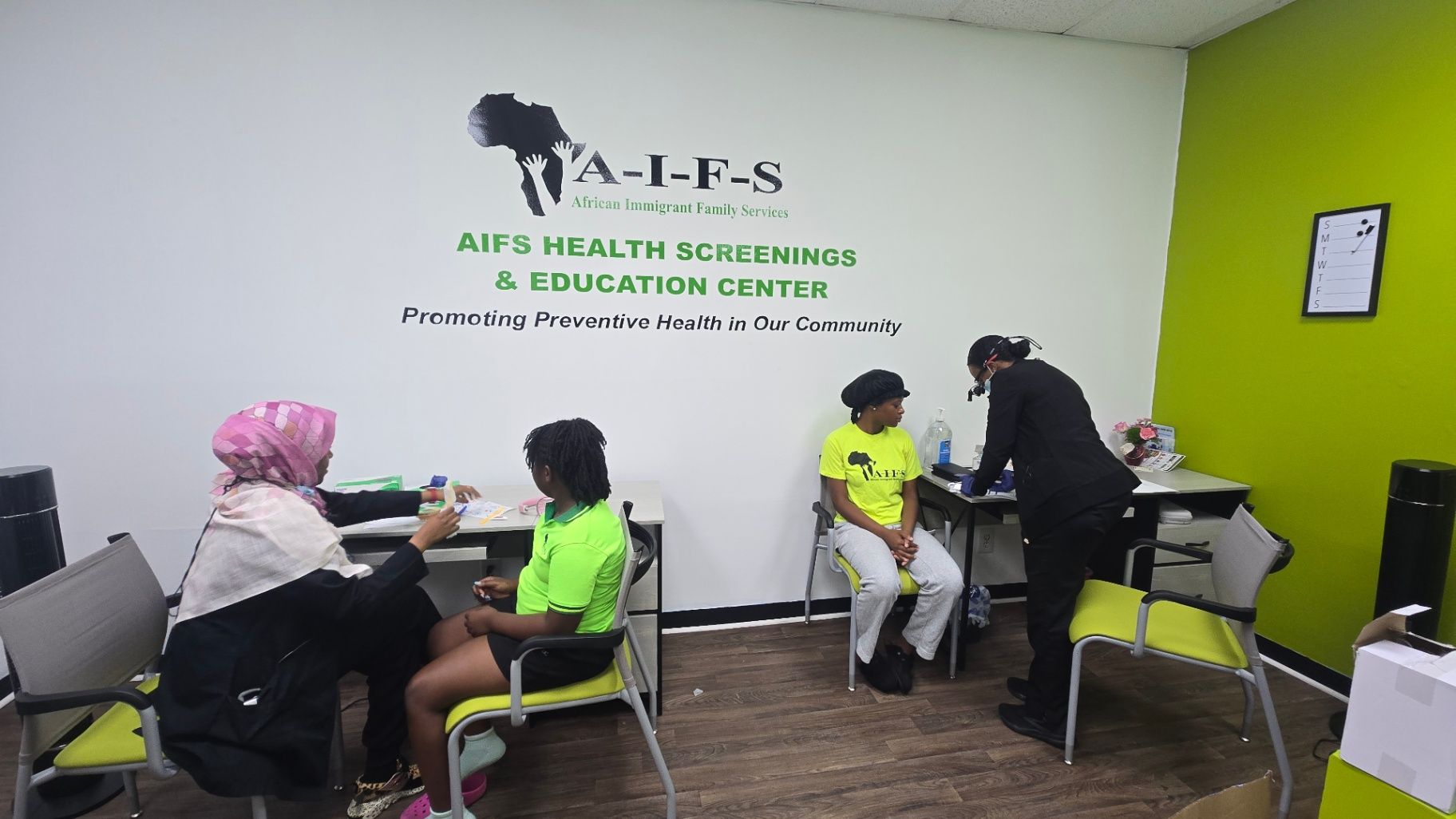 A room with two tables where adults sit during a screening activity beneath an AIFS Health Screenings and Education Center wall sign.