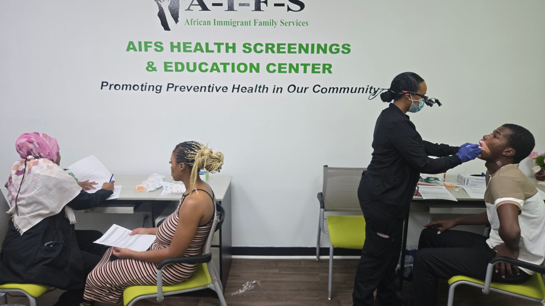 A masked healthcare worker examines a seated person while another adult holds paperwork in a room with AIFS signage.