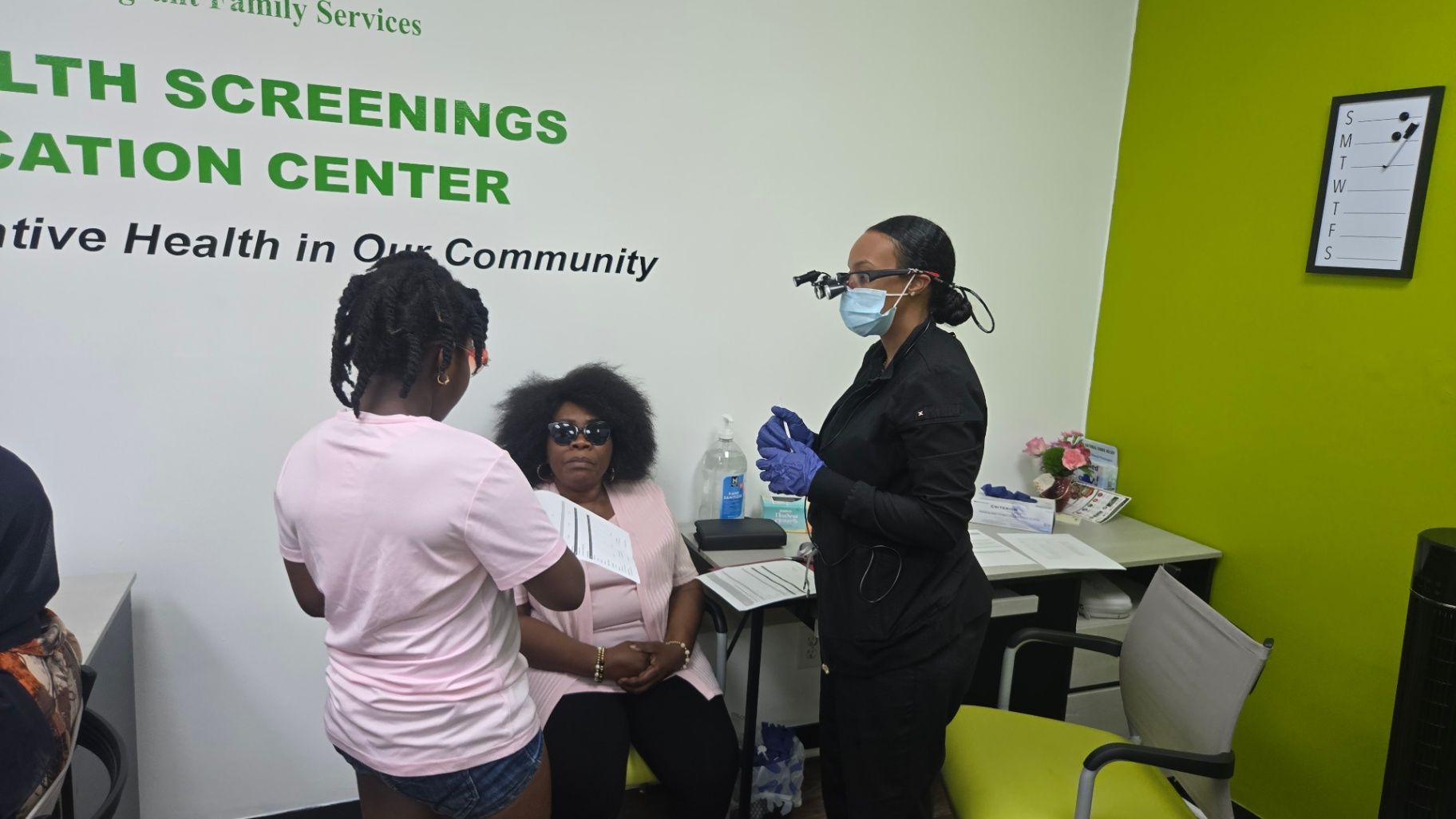 A masked healthcare worker stands beside a seated adult while another person holds paperwork in a room with green walls and AIFS signage.